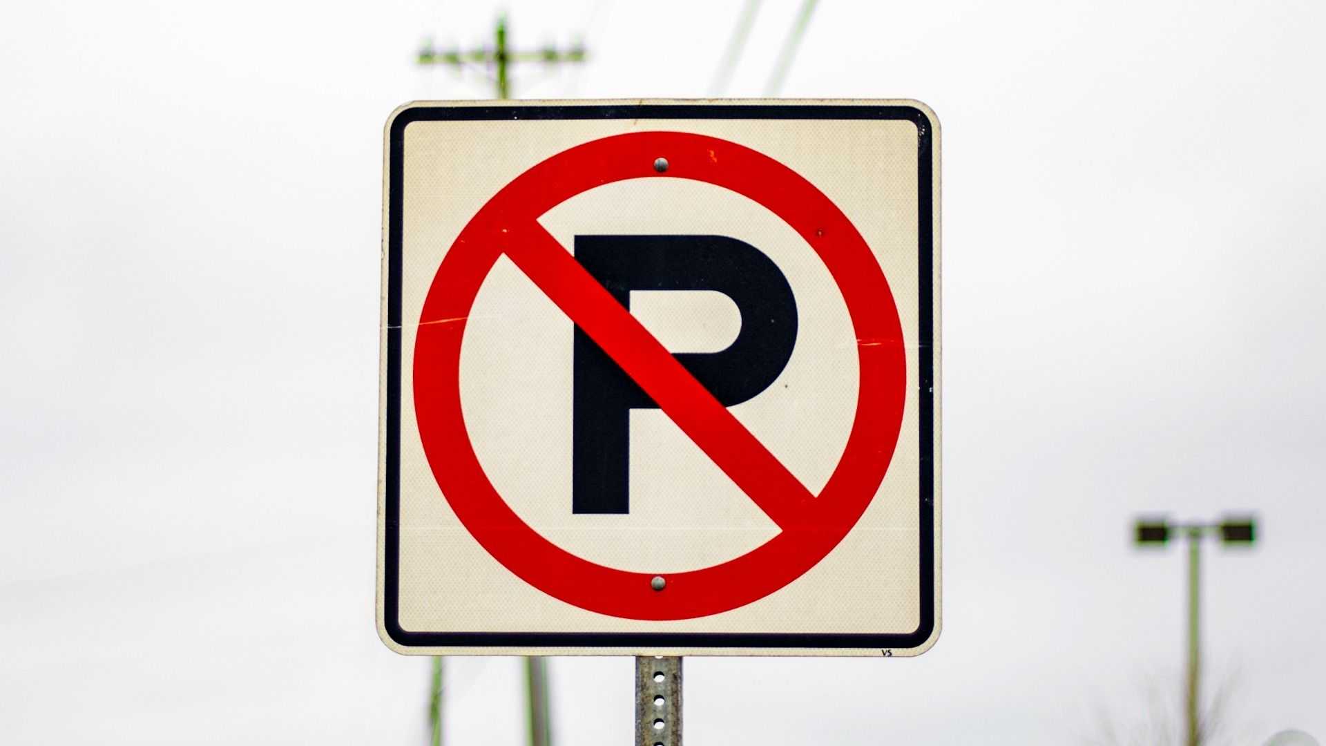 No parking sign featuring a black letter “P” crossed out by a red circle and diagonal slash, mounted on a metal post with utility poles blurred in the background.